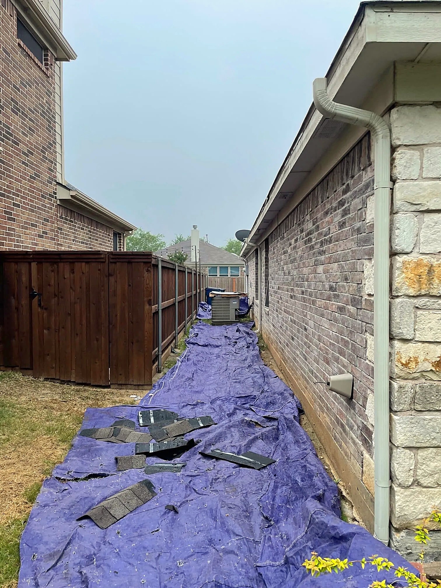 A blue tarp laid along the side yard of a brick house to catch debris